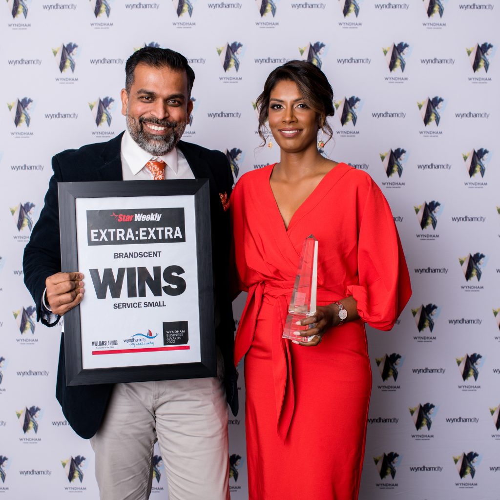 BrandScent co-founders celebrating their win at the Wyndham City Business Awards, holding trophies for Best Small Service Business with a branded media wall in the background