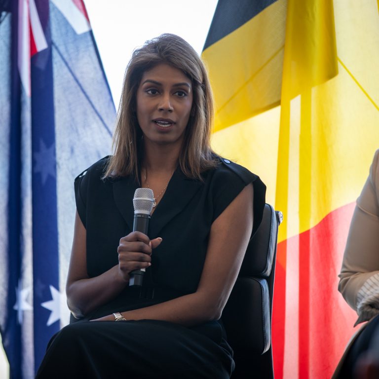 Woman speaking into a microphone during a panel discussion, seated beside another professional with Australian and Aboriginal flags in the background at a formal event