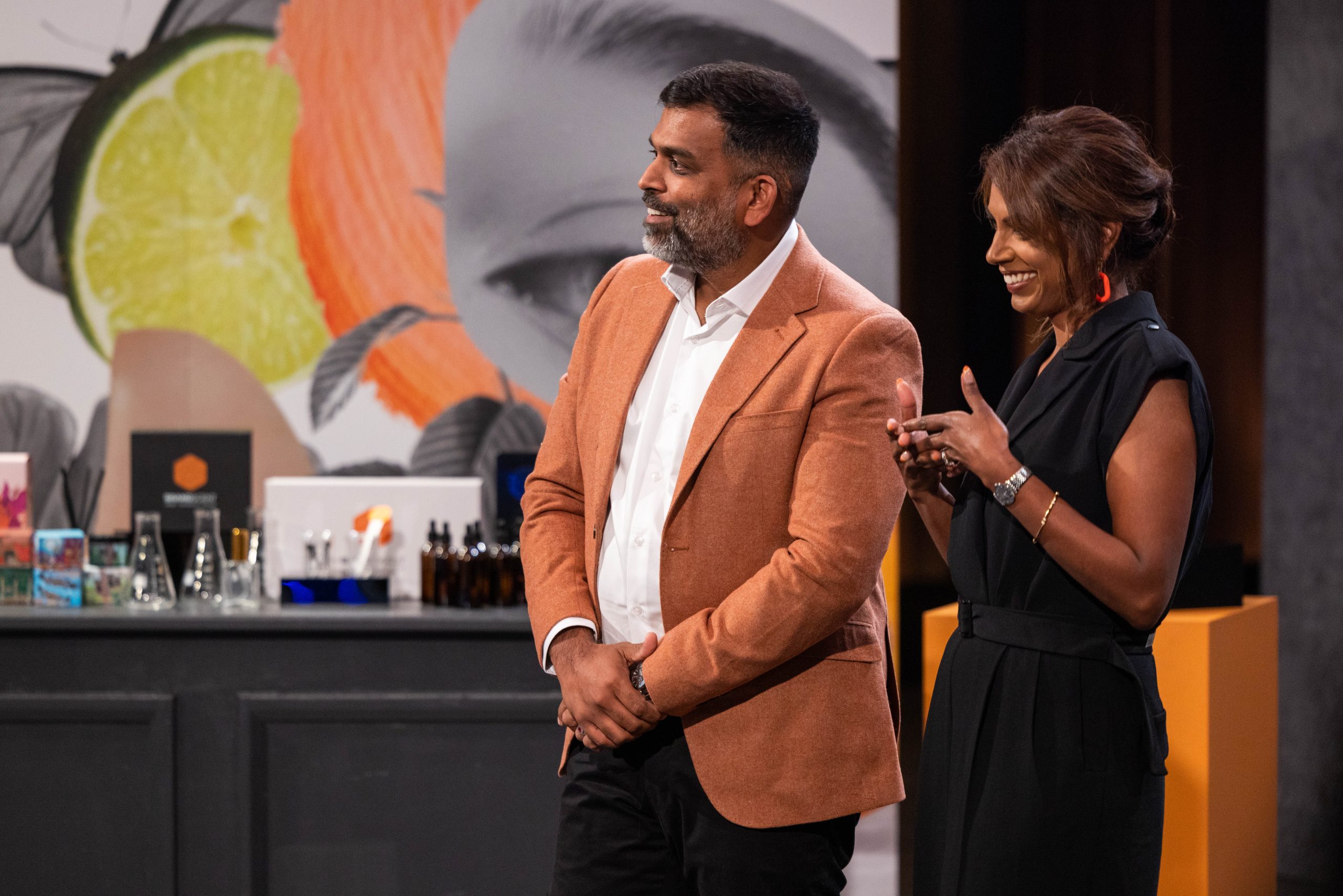 Entrepreneurial duo presenting their product line on a TV show set, standing confidently in front of a display of cosmetics and packaging with vibrant backdrop art.