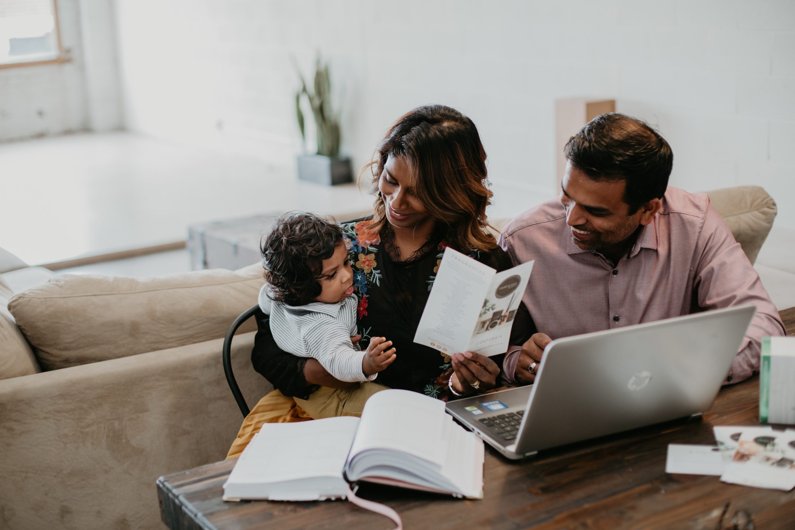 Young family working together at home, with a mother holding a child while reviewing documents and the father smiling beside a laptop on the table.