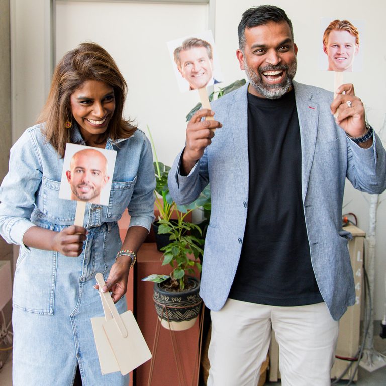 Two people laughing and holding cutout face props during a fun office or team-building activity, standing in a modern indoor space with potted plants