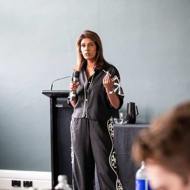 Confident woman delivering a presentation while holding notes, standing in front of a dark green wall and black-covered table in a professional setting