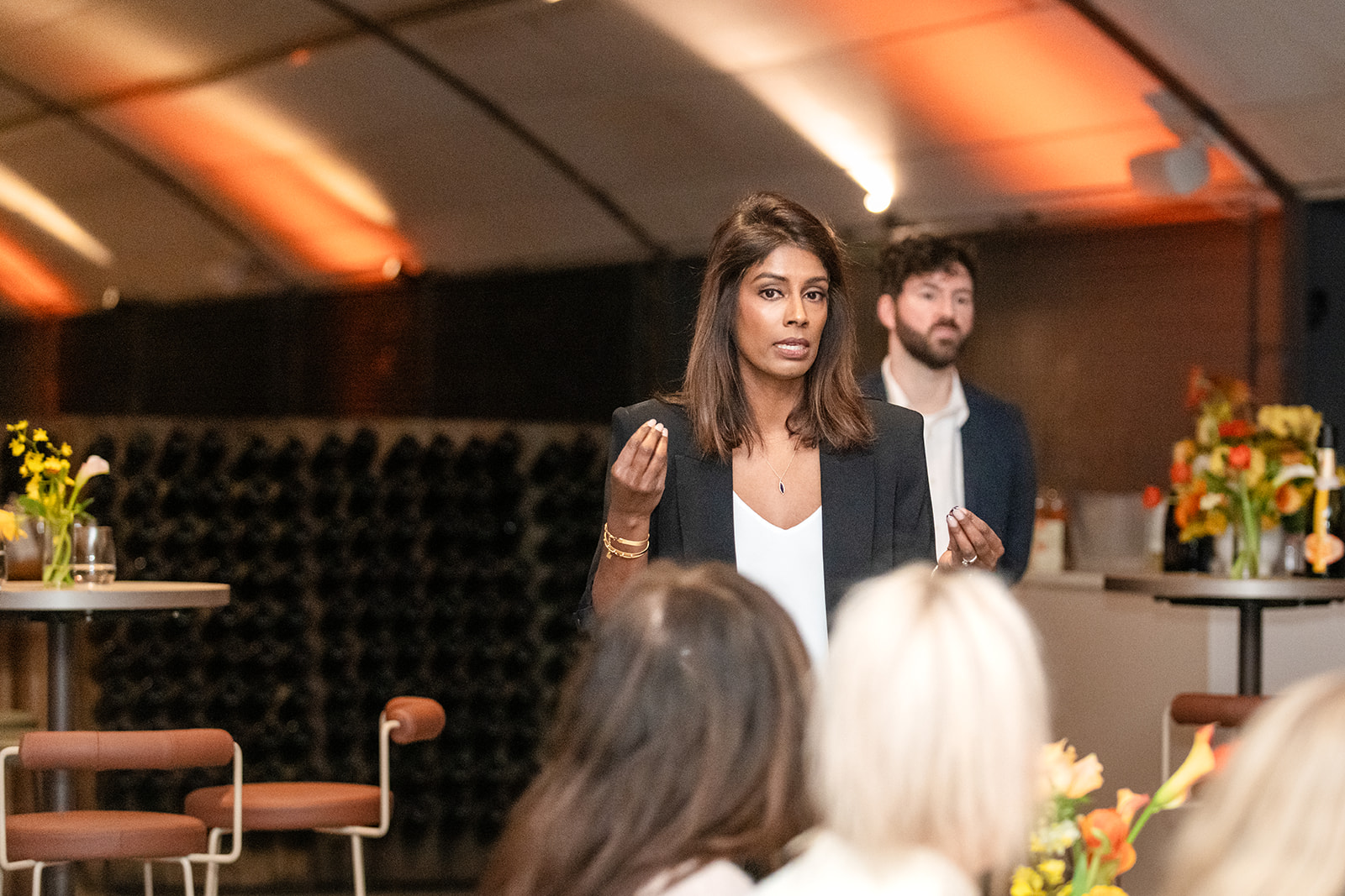 Woman smiling and engaging with guests at a wine cellar event, standing beside a table with flowers and a drinks menu, with wine bottles stacked in the background.