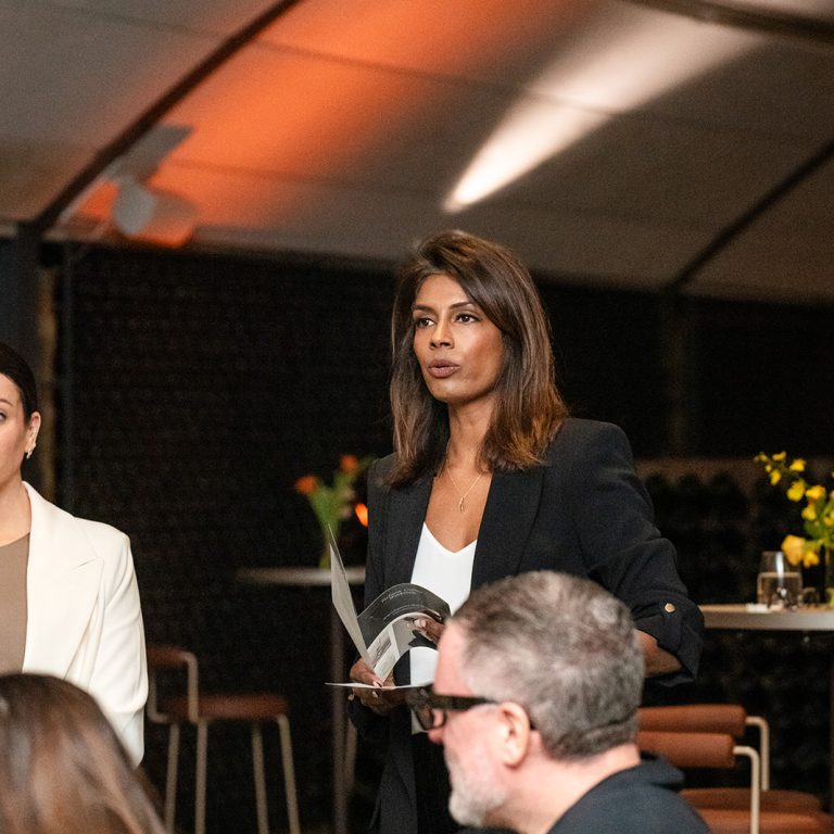 Woman smiling and engaging with guests at a wine cellar event, standing beside a table with flowers and a drinks menu, with wine bottles stacked in the background.