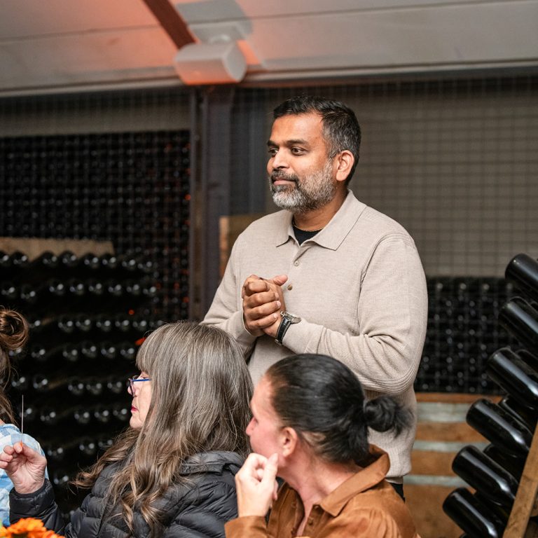 Man standing and speaking at a wine cellar dinner event, surrounded by seated guests and floral table arrangements, with stacked wine bottles in the background.
