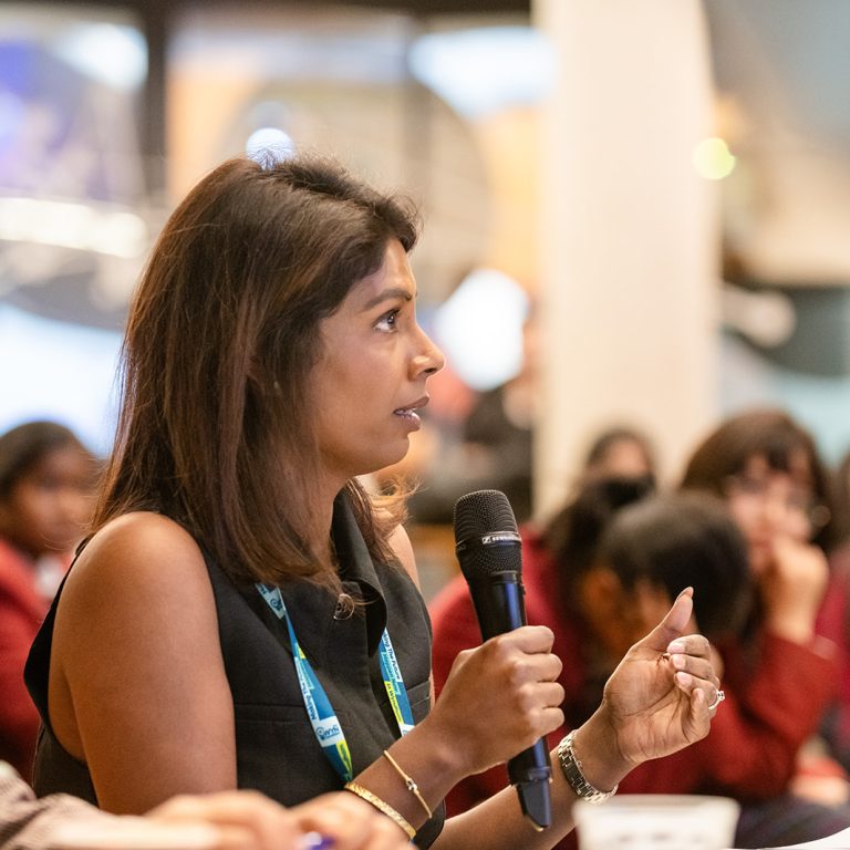 Woman speaking into a microphone at a professional conference panel, engaging attentively in front of a diverse seated audience